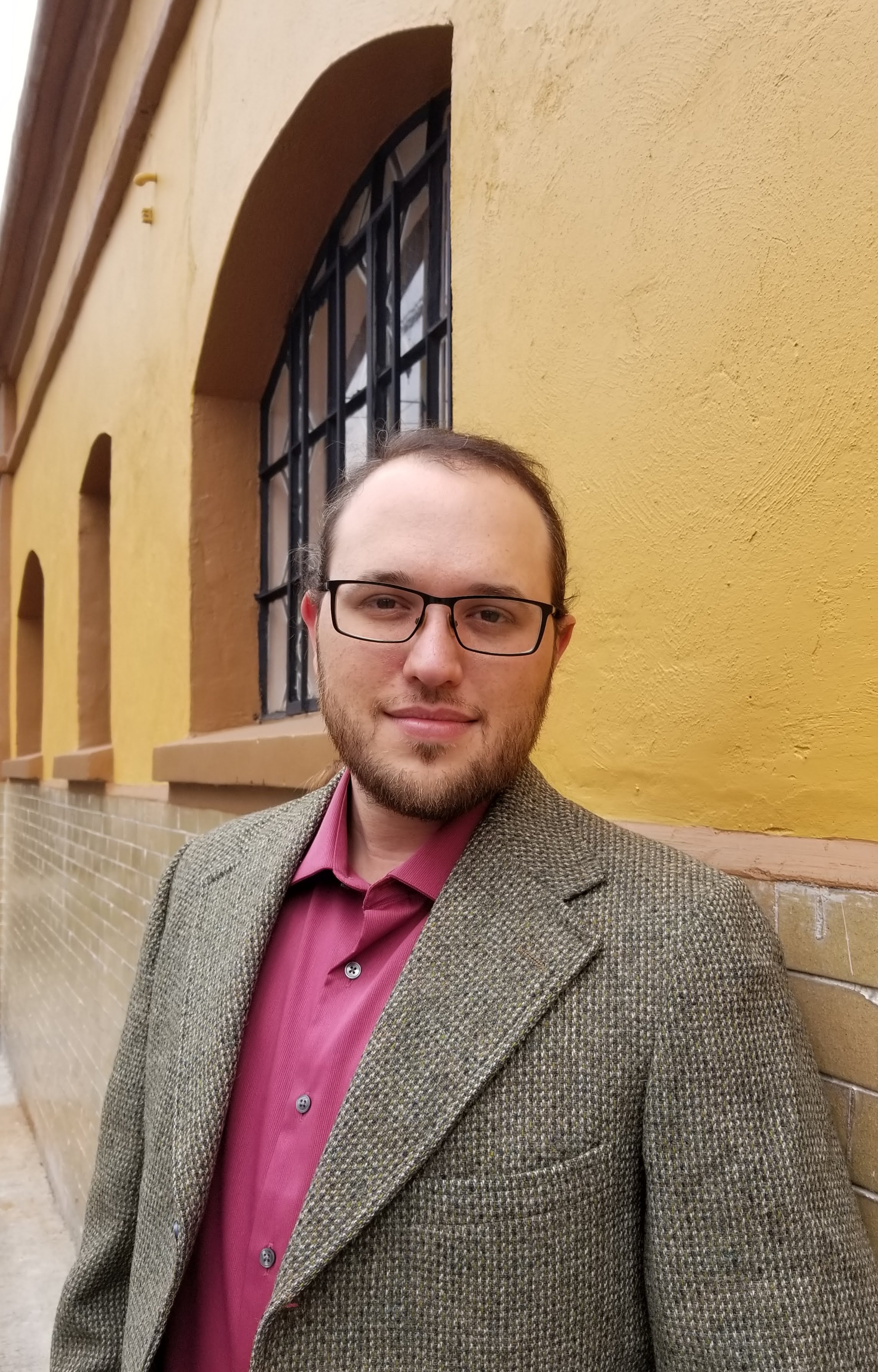 man in suit jacket and button-down shirt with glasses standing in front of a yellow building