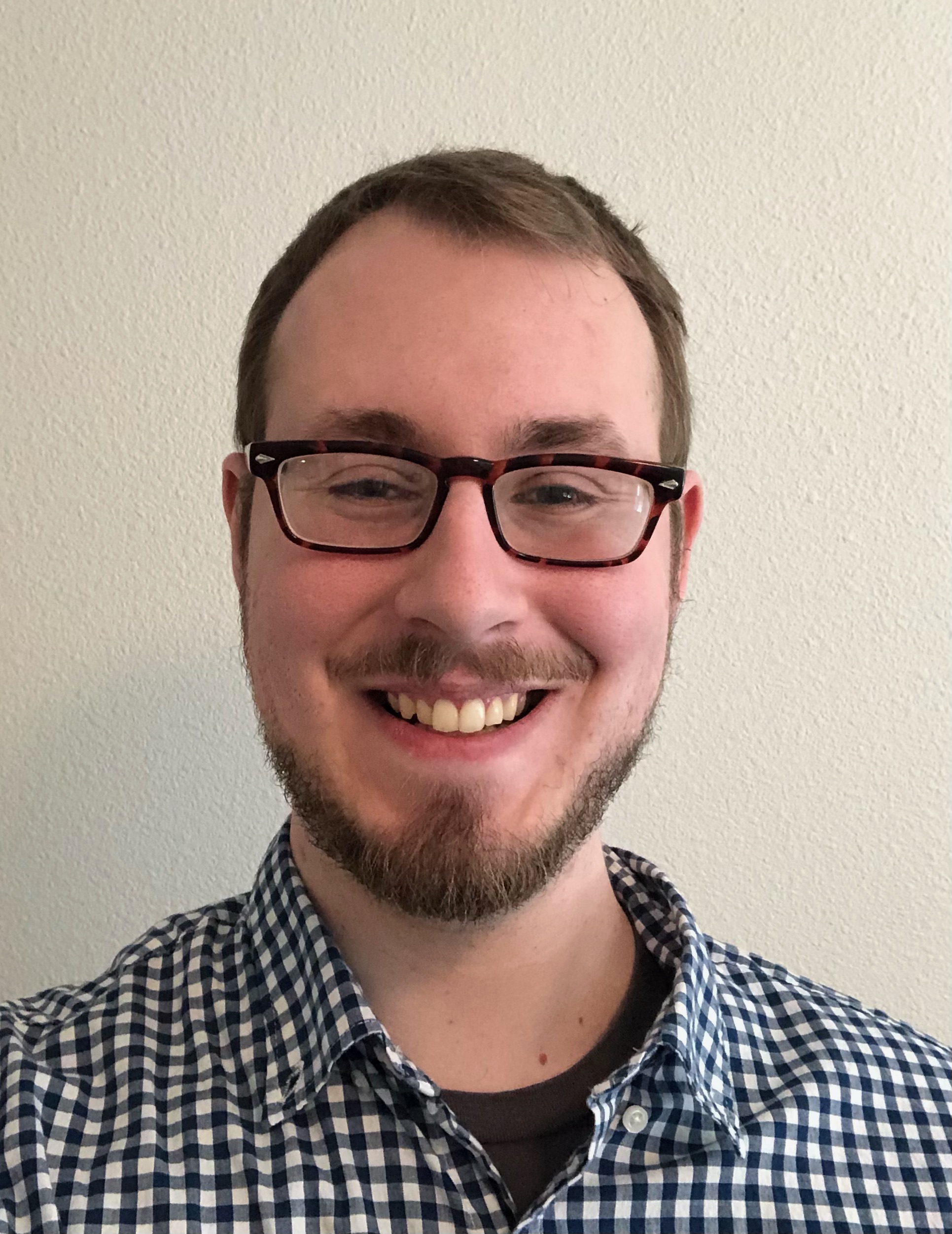 headshot of a man with short hair and glasses wearing a plaid shirt in front of a blank wall