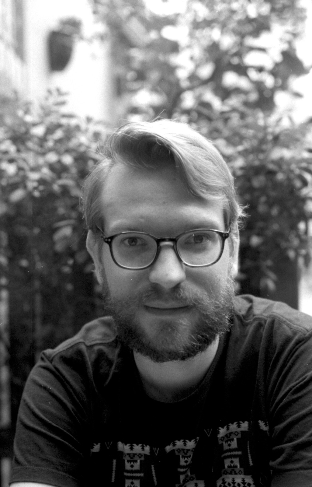 black and white photo of a man wearing glasses. He is outside in front of bushes.