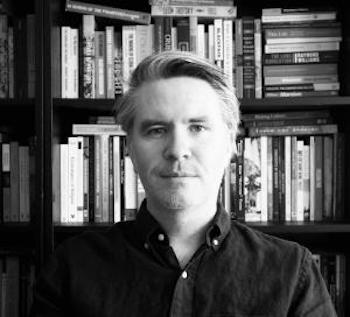 black and white photo of a man in a dark shirt sitting in front of bookshelves