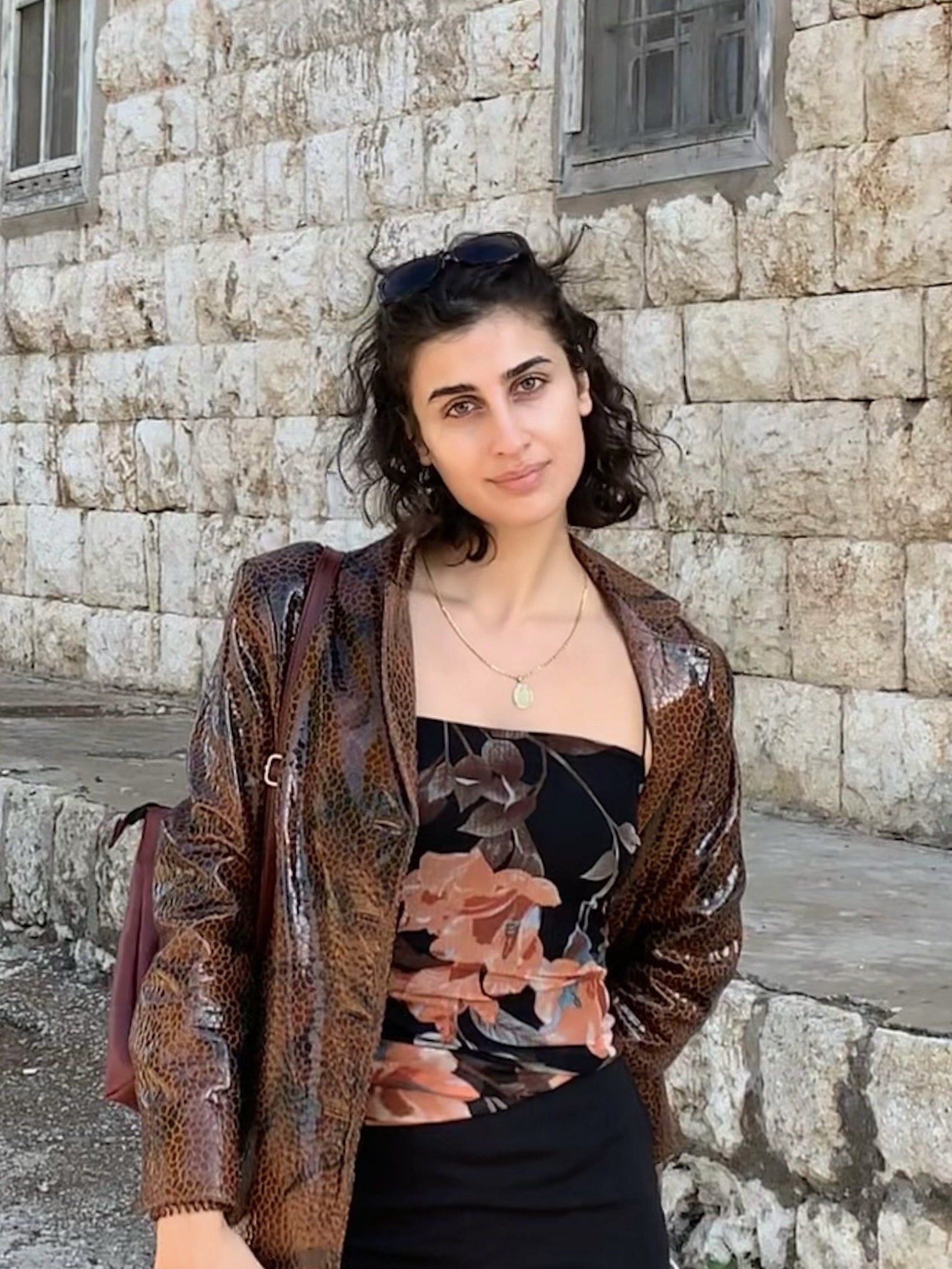 woman with short, curly hair in a leather jacket and black top standing in front of a stone wall