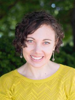 Headshot of a woman with short, curly hair wearing a yellow sweater with trees in the background